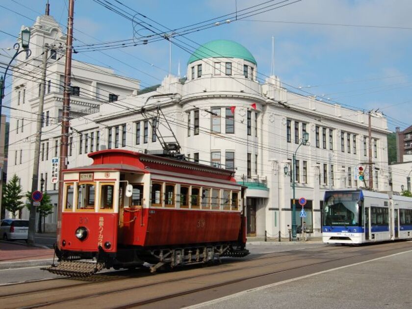 大正時代に造られた建造物の前を行く復元チンチン電車「箱館ハイカラ號」と、スムーズに乗り降りできる超低床電車「らっくる号」